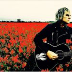 Mike Peters in a flower field with acoustic guitar.