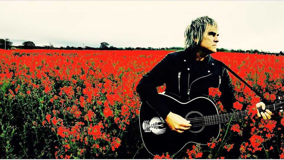 Mike Peters in a flower field with acoustic guitar.
